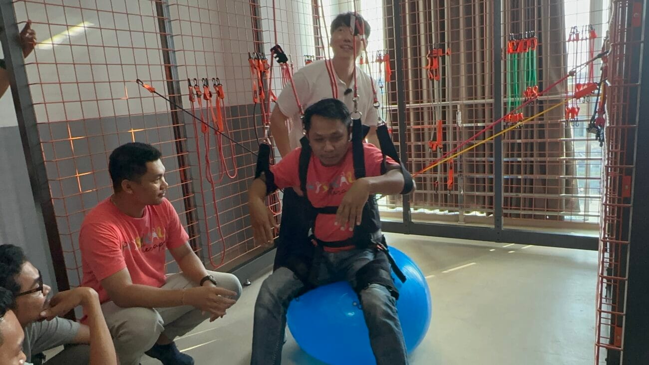 Seorang peserta terapi melakukan latihan keseimbangan menggunakan Wall Sling System dan gym ball dengan pendampingan fisioterapis.